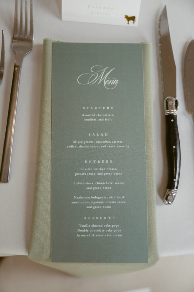 A close-up of a wedding reception place setting featuring a sage green napkin with a printed menu placed on top. The menu lists starters, salad, entrées, and desserts, and is surrounded by silverware on a white tablecloth.