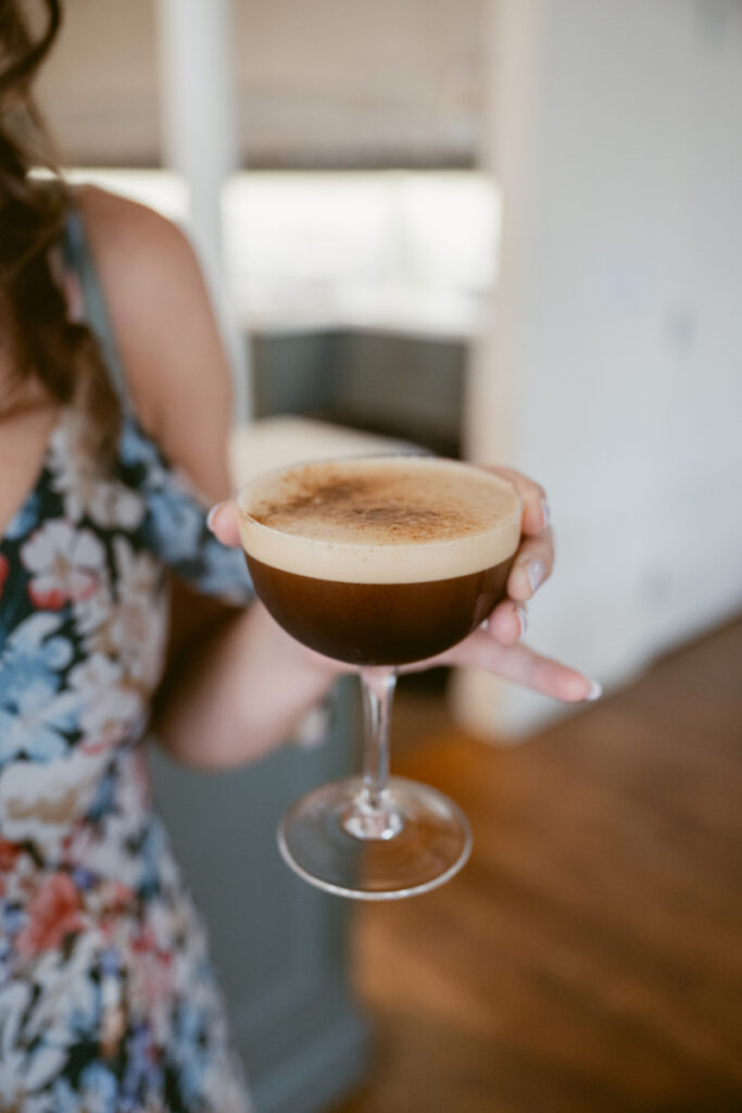 A woman in a floral dress holds an espresso martini in a coupe glass, with the creamy foam and dusting on top clearly visible. The background is softly blurred, drawing focus to the drink.