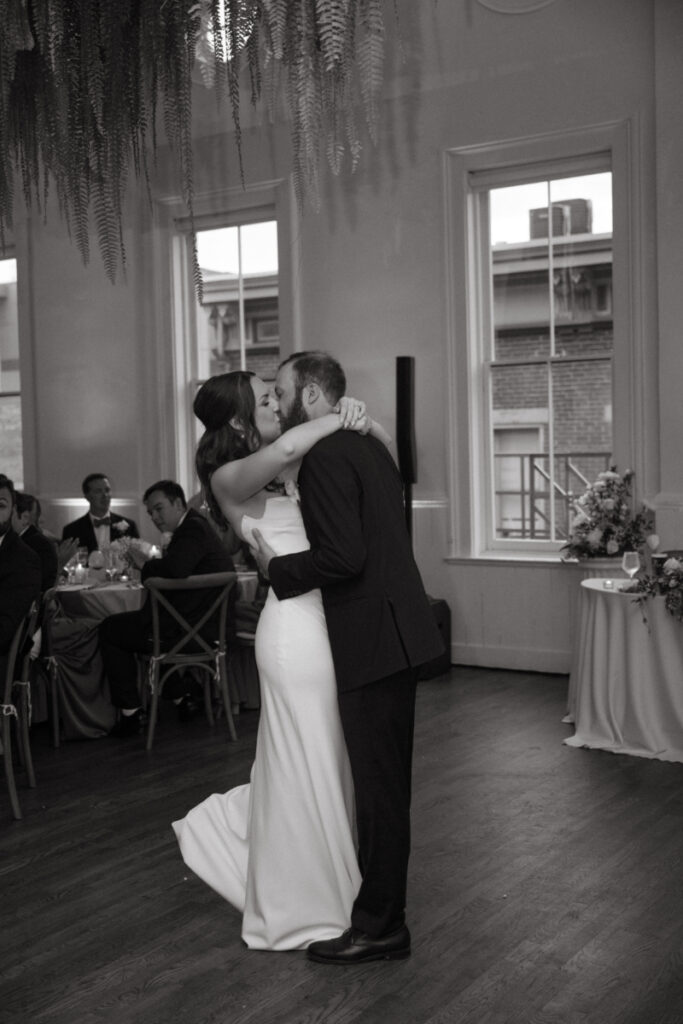 A bride and groom share a kiss during their first dance in a warmly lit reception space with large windows and hanging greenery overhead.