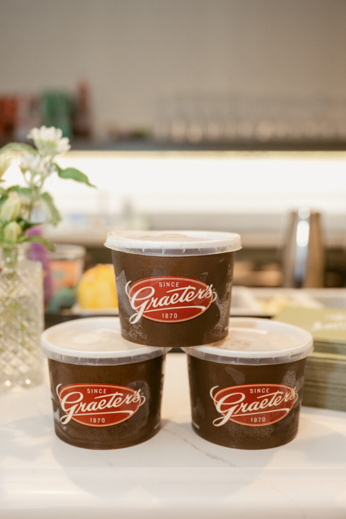 Three containers of Graeter's Ice Cream stacked on a counter, with a floral arrangement and bar elements in the background. The containers are dark brown with red and white labels.