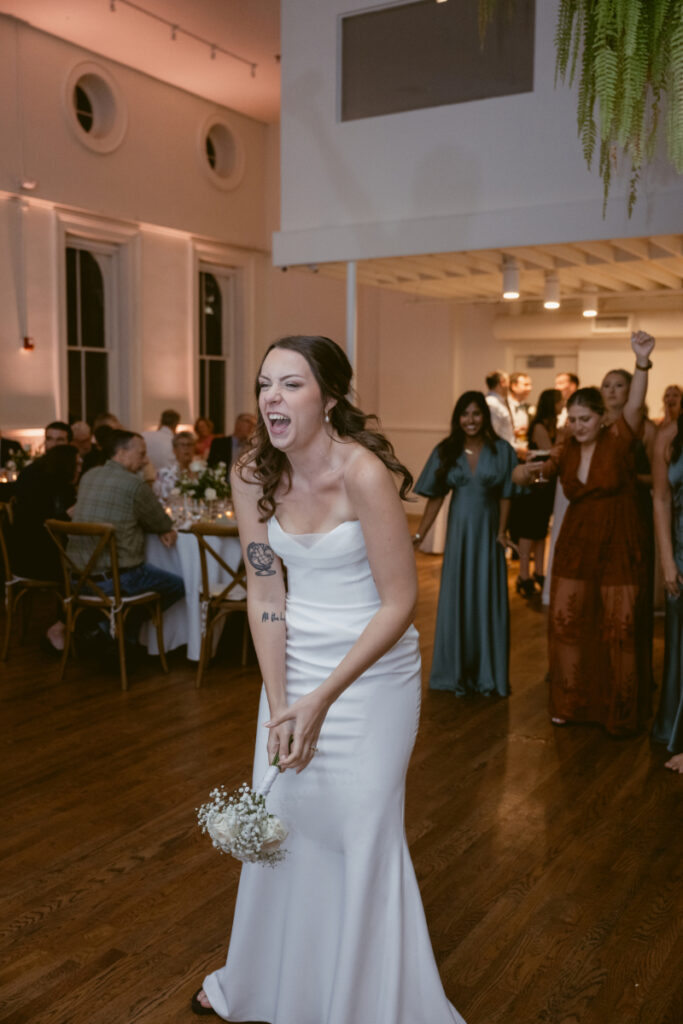 The bride laughs while holding her bouquet, preparing to toss it as guests eagerly wait behind her at the reception.