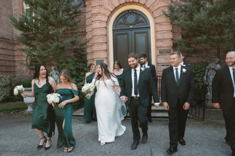Bride and groom walk hand in hand with wedding party outside Kingwood Hall at Ohio garden wedding.