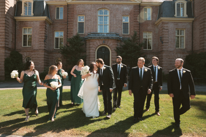 Bride and groom kiss while walking with bridal party in front of mansion at their Ohio garden wedding.