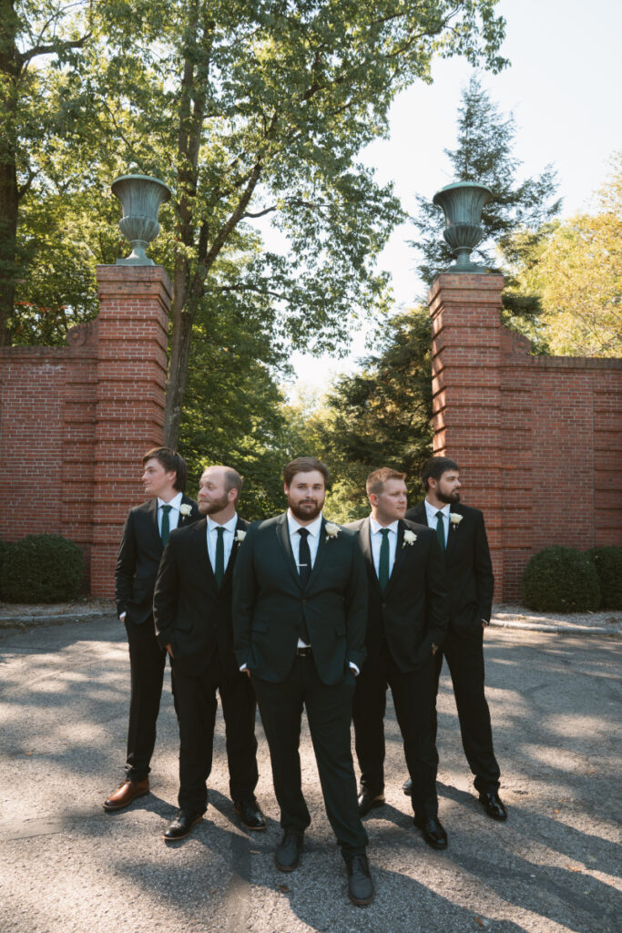 Groom and groomsmen pose confidently near brick gate at sunlit Ohio garden wedding.