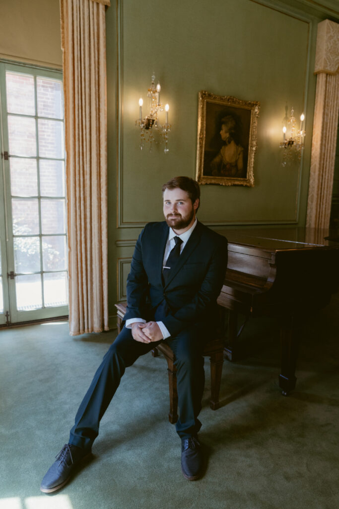 Groom sits by piano inside historic mansion before Ohio garden wedding ceremony.