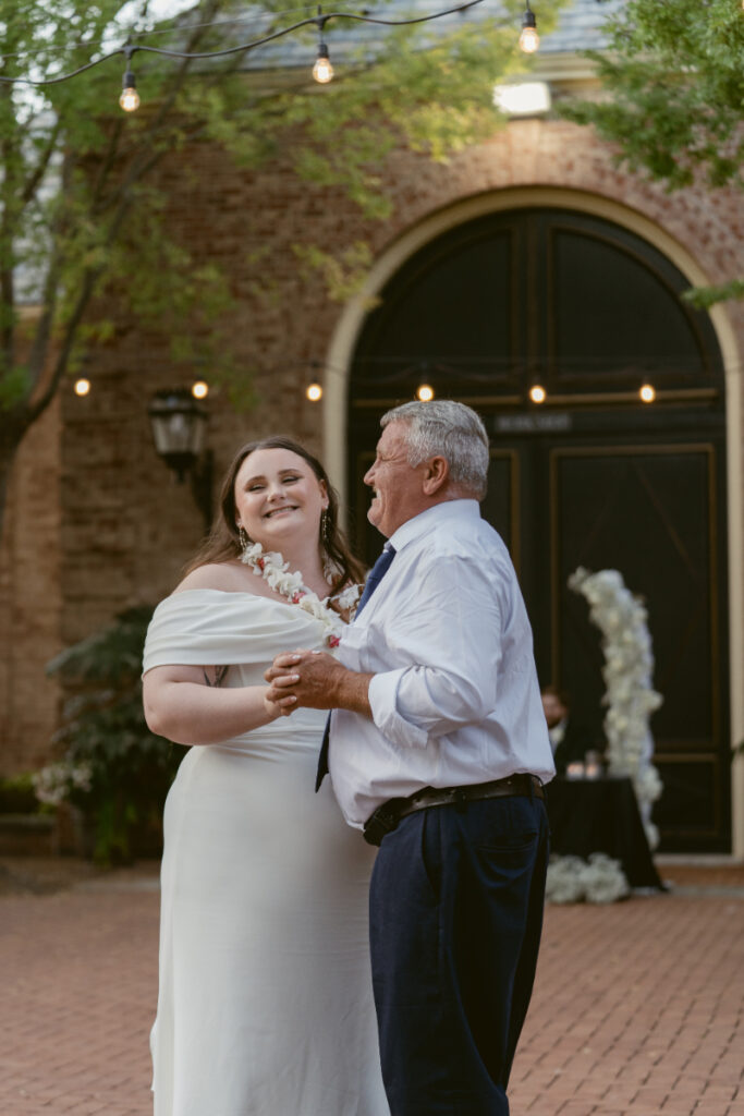 Bride smiles while sharing a sweet father-daughter dance under string lights in the courtyard.