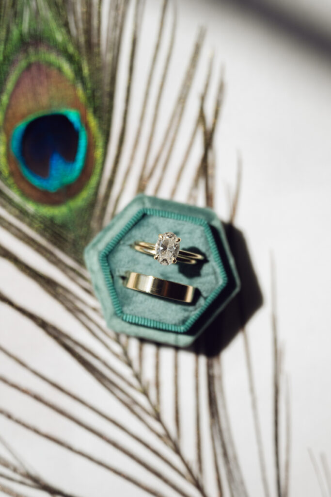 Close-up of wedding rings in a teal velvet box beside a vibrant peacock feather.