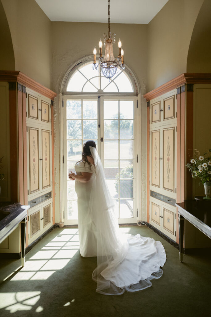 Bride in gown and veil stands in sunlit hallway
