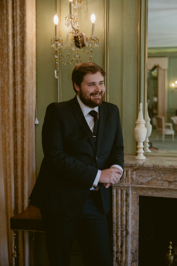 Groom smiling and leaning on fireplace mantel before Ohio garden wedding ceremony.