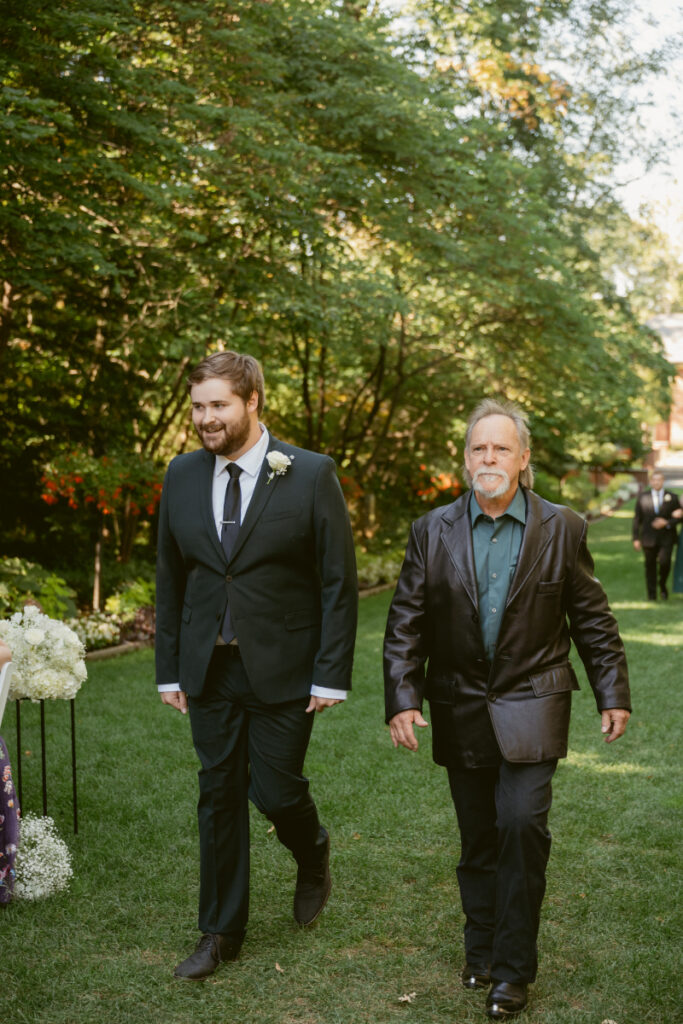 Groom walks down grassy aisle with family member during outdoor Ohio garden wedding ceremony.