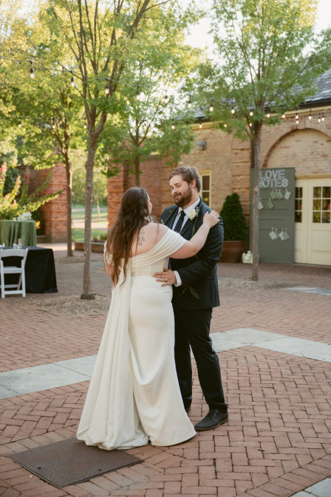 Bride and groom share a sweet first dance in a brick courtyard surrounded by trees and sunlight.