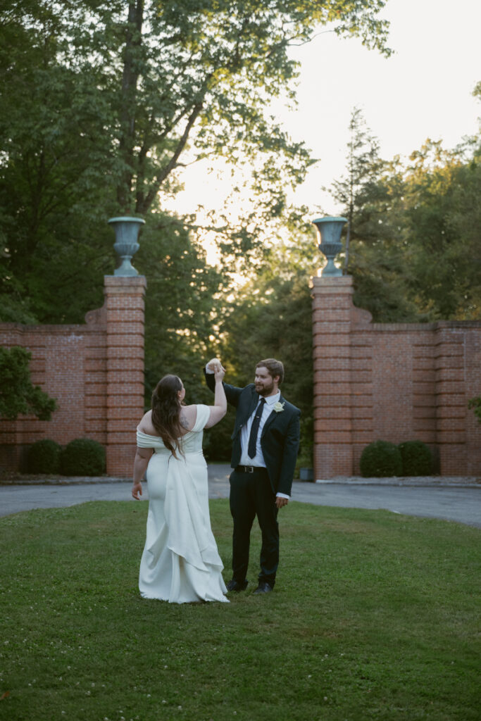 Groom twirls the bride during golden hour in a garden courtyard surrounded by brick pillars.