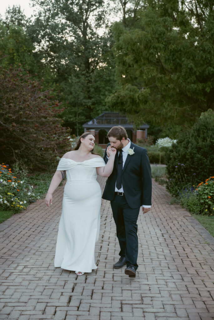 Bride and groom walk a brick path through lush gardens as the groom kisses her hand.