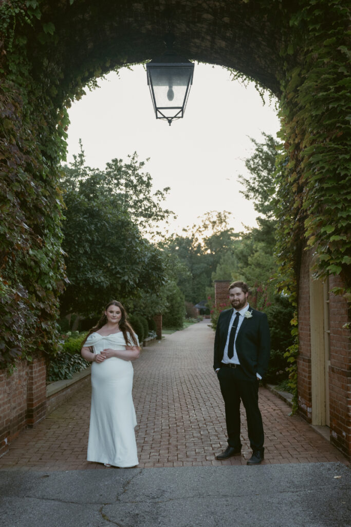 Bride and groom stand under an ivy-covered archway at sunset on a brick garden path.