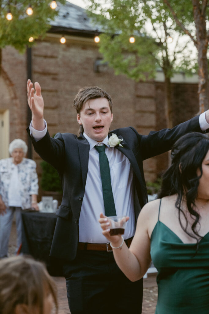 Wedding guest dancing with arms raised during outdoor reception at sunset with string lights overhead.