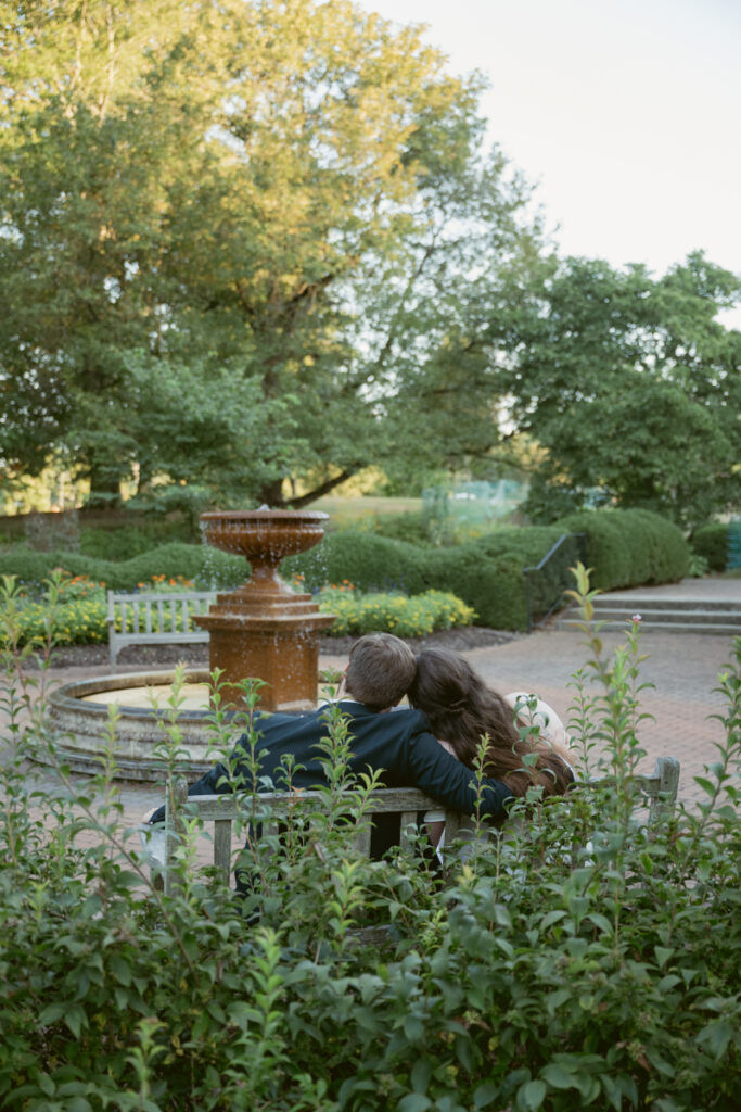 Bride and groom share a quiet moment on a bench beside a fountain at sunset.