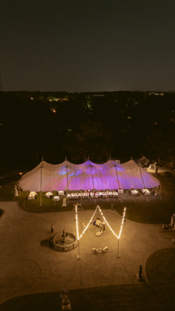 Outdoor wedding tent lit with purple lights at night, surrounded by string lights and guests.