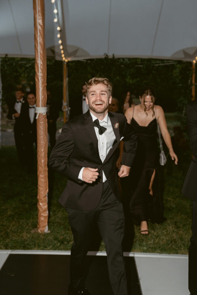 Groomsman in tuxedo joyfully runs onto the dance floor under a tent at night.