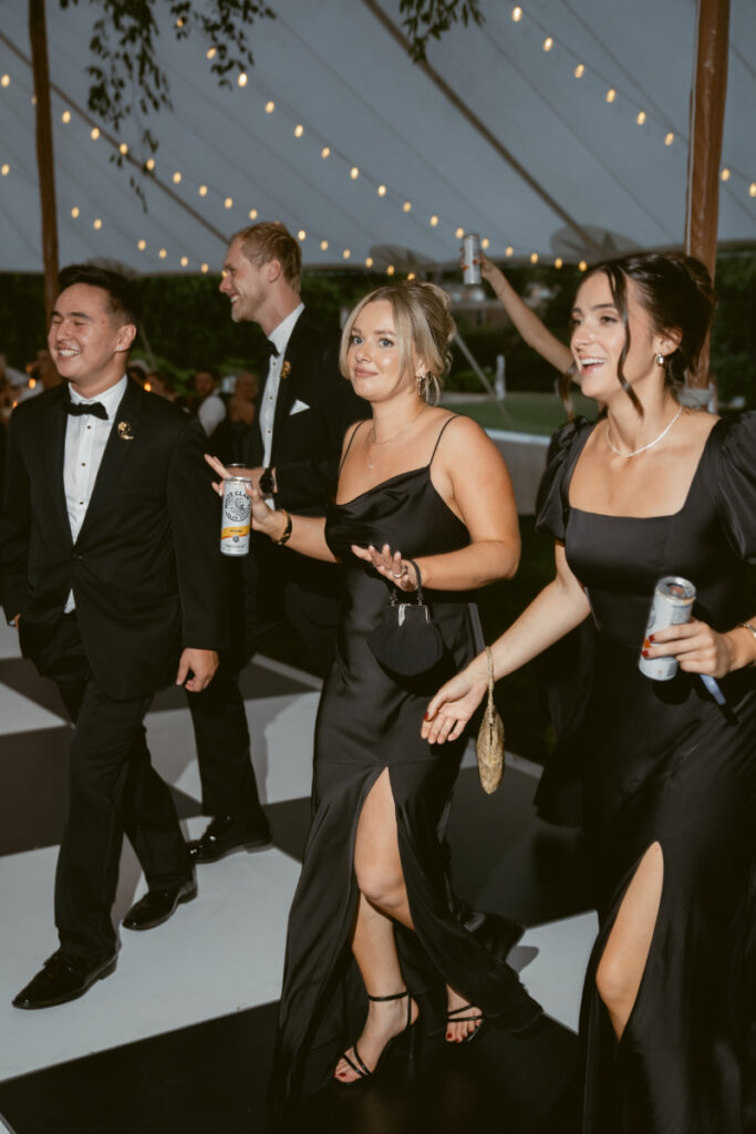 Bridesmaids and groomsmen in formal attire enter the reception tent smiling and holding drinks.