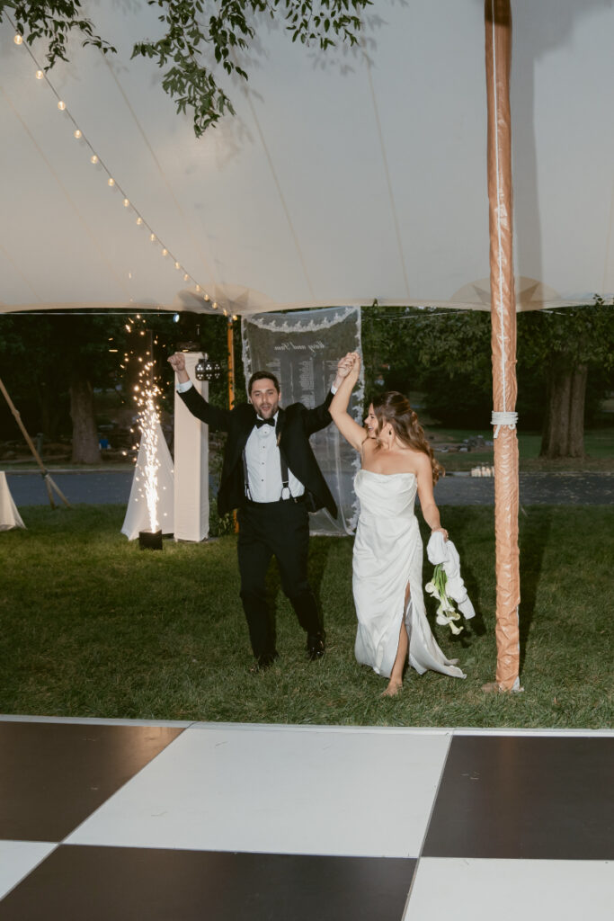 Bride and groom joyfully enter the reception tent hand in hand with sparklers behind them.