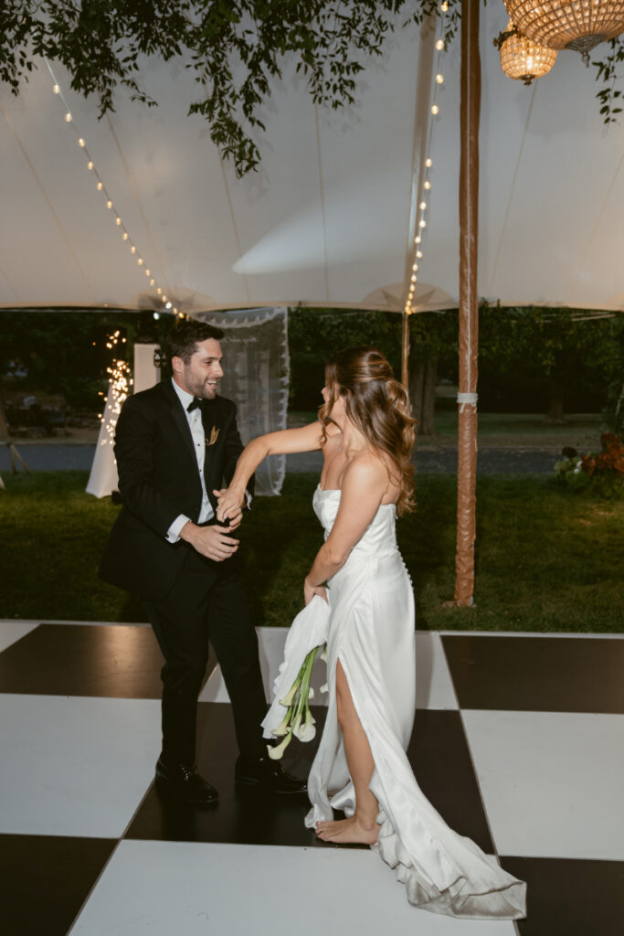 Newlywed couple dances joyfully on a black and white checkered floor under a tent.
