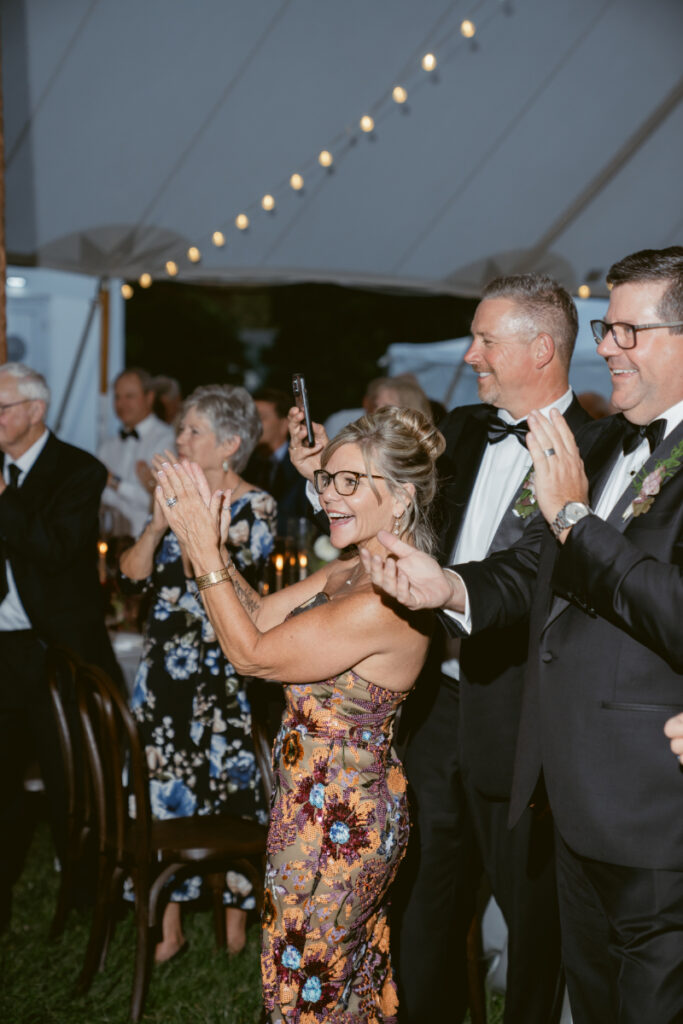 Guests cheer and applaud under a tent during an outdoor evening celebration.