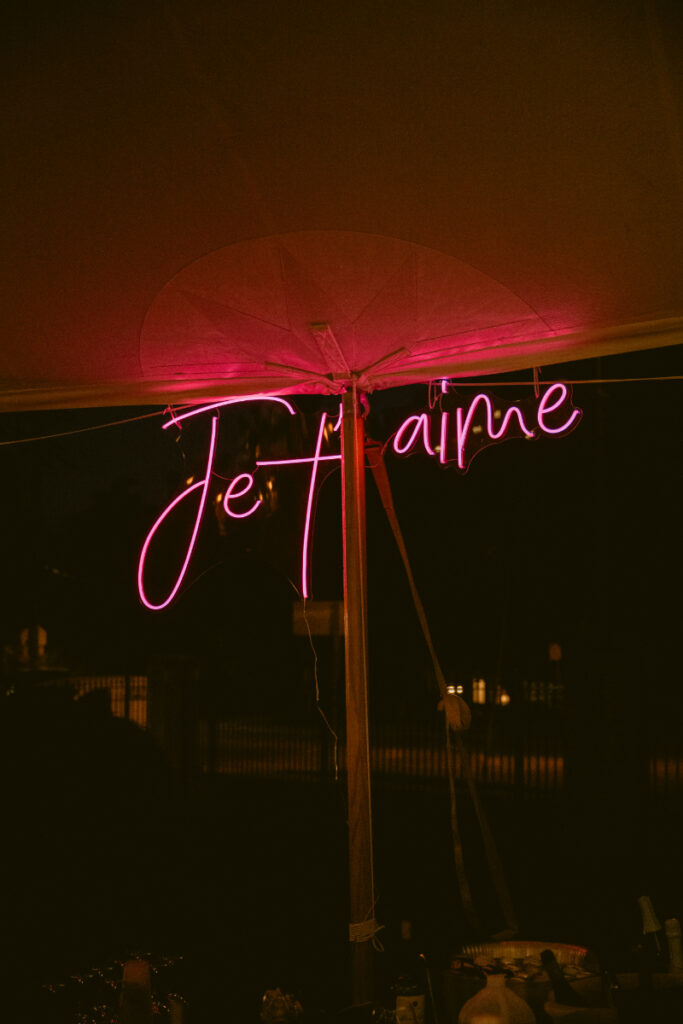 Pink neon "Je t’aime" sign glows under a tent during an evening wedding celebration.
