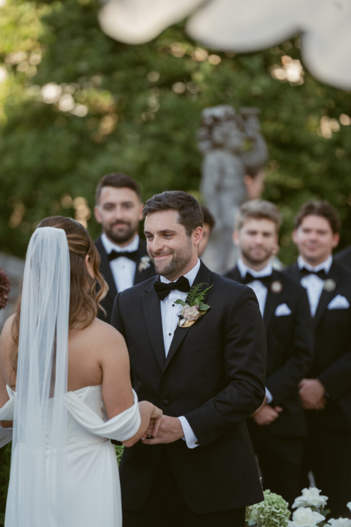 Groom smiling at bride during outdoor wedding ceremony, surrounded by groomsmen in black tuxedos.