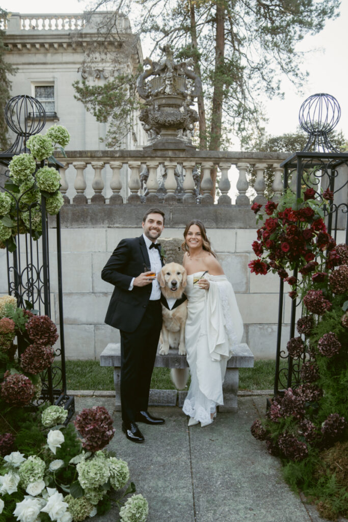 Bride and groom pose with their dog in elegant wedding attire, surrounded by lush floral arrangements.