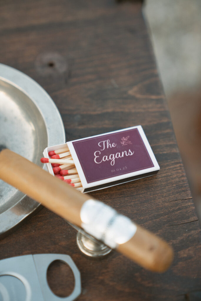 Close-up of personalized matchbox and cigar on a wooden table at an outdoor wedding celebration.