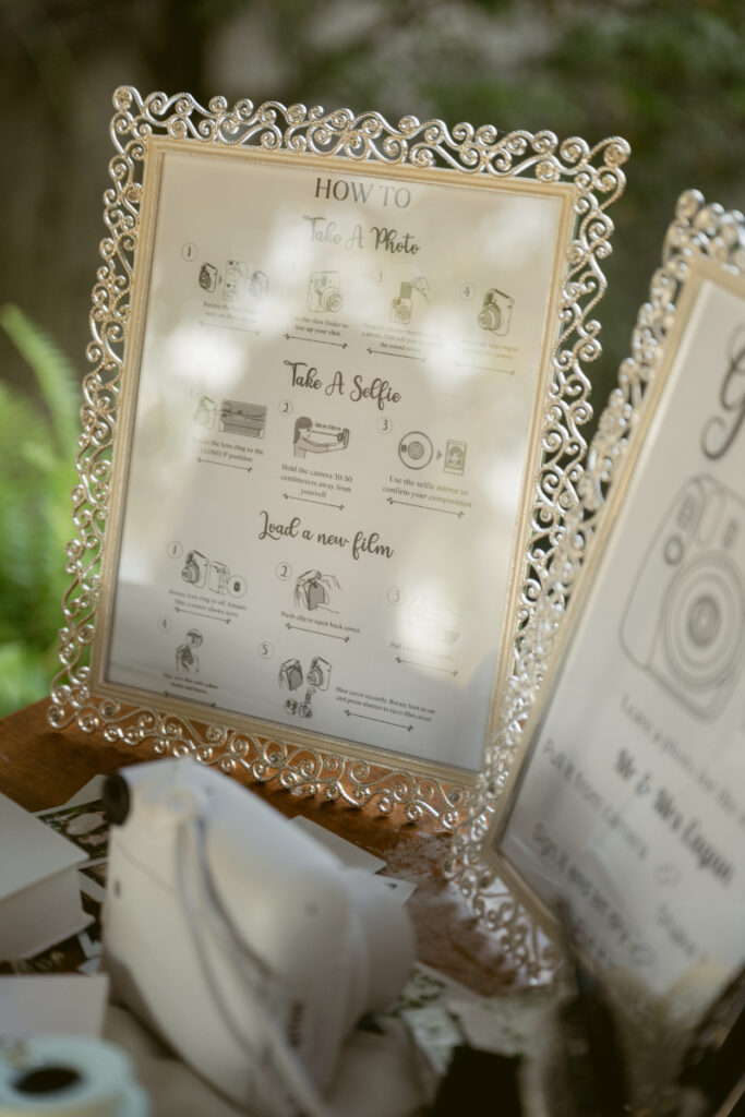 Elegant framed sign with photo booth instructions displayed beside an instant camera at a wedding.