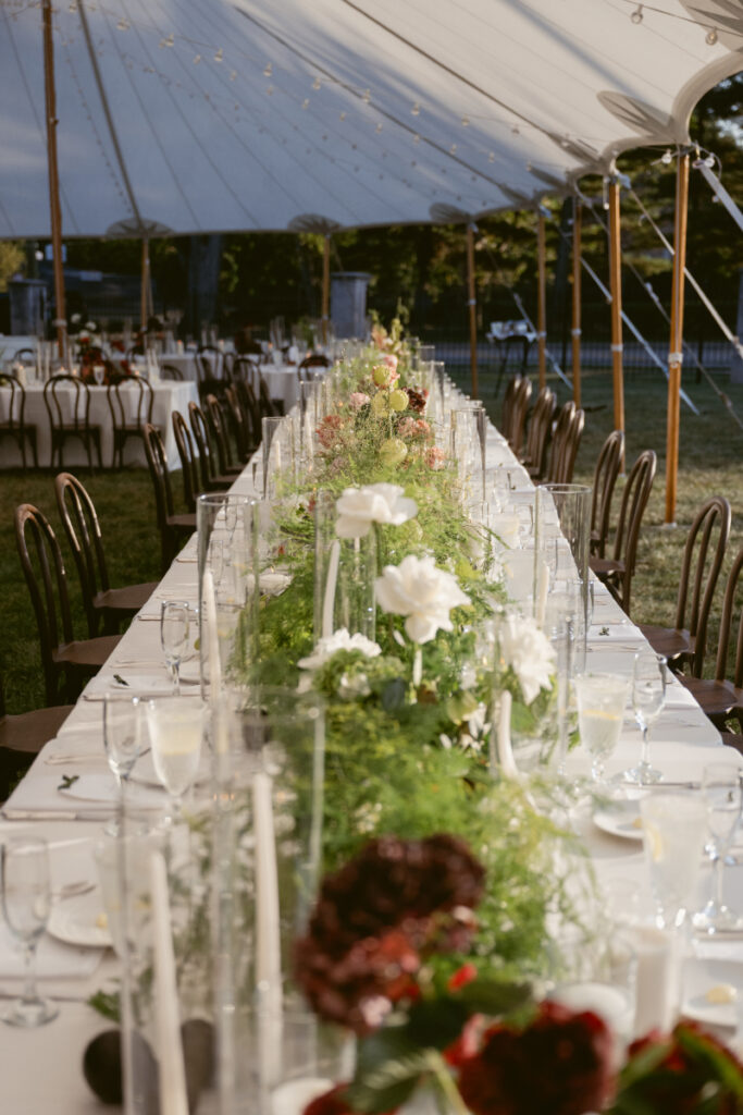 Long outdoor reception table with elegant floral centerpiece, surrounded by chairs under a large tent.