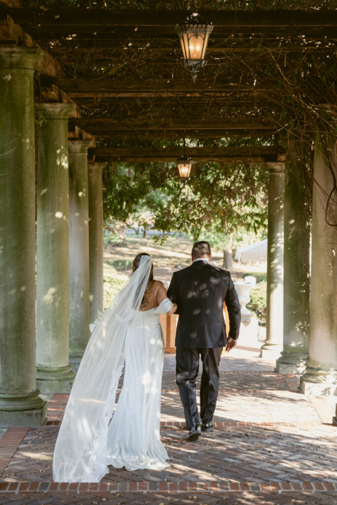 Bride walks arm in arm with father under vine-covered stone pergola toward wedding ceremony location.
