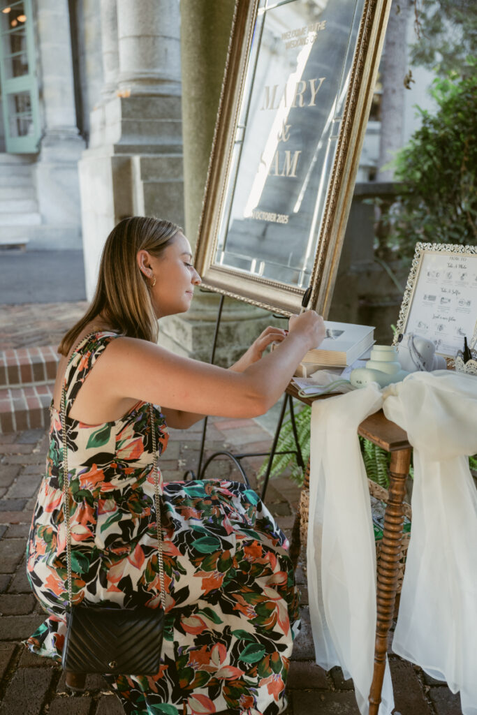 Guest in floral dress uses instant camera at wedding photo guestbook station with elegant signage.