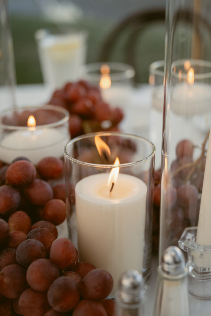 a glowing white candle in a glass holder with red grapes surrounding it