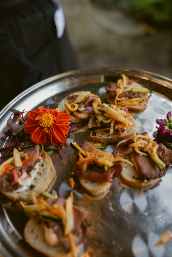Gourmet appetizers on toasted bread served on silver tray with bright floral garnish at event.
