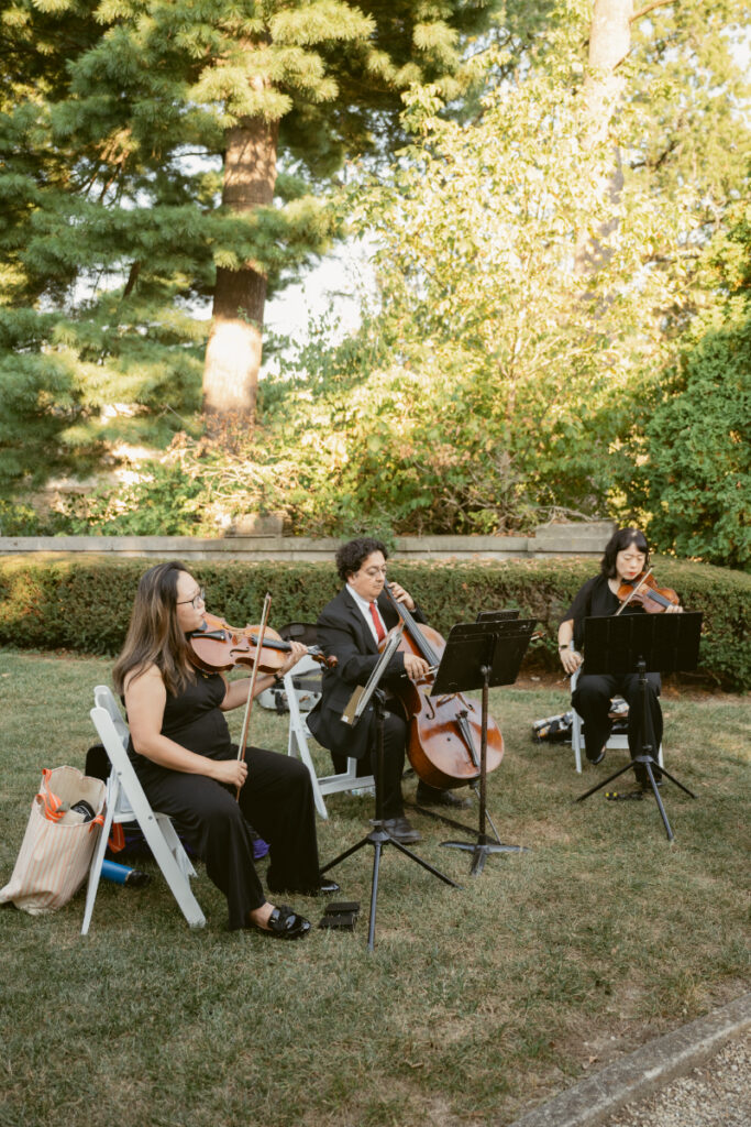 String trio performing outdoors on a lawn during a formal event, surrounded by trees and sunlight.