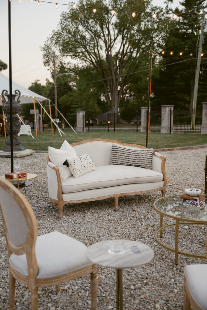 Elegant outdoor wedding lounge setup with vintage sofa, chairs, and string lights at sunset.