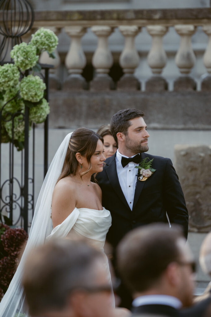 Bride and groom standing together during outdoor ceremony, both looking off into the distance smiling.