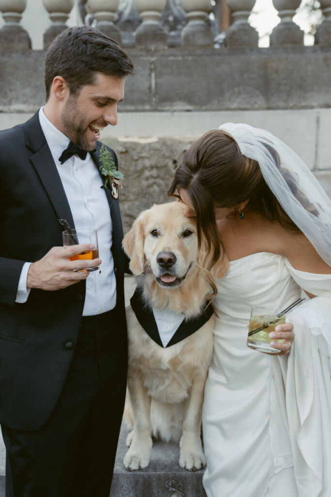 Bride and groom smiling with their golden retriever in a tuxedo at elegant outdoor wedding.