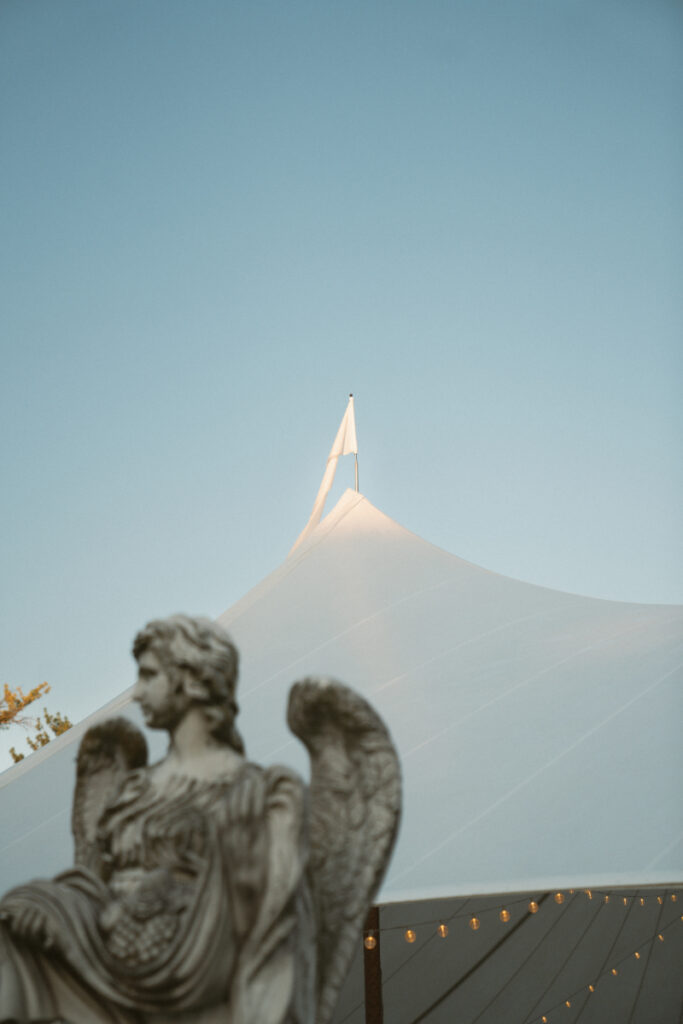 White tent peak under blue sky with angel statue and string lights in the foreground.