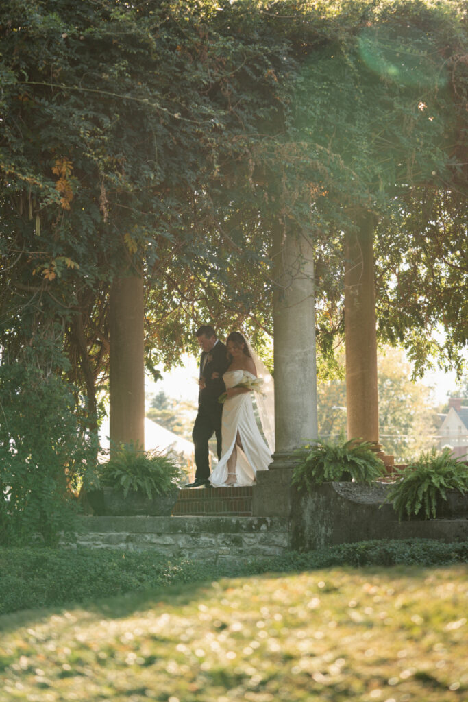 Bride walks with her escort through a sun-dappled garden path, framed by tall columns and lush greenery.