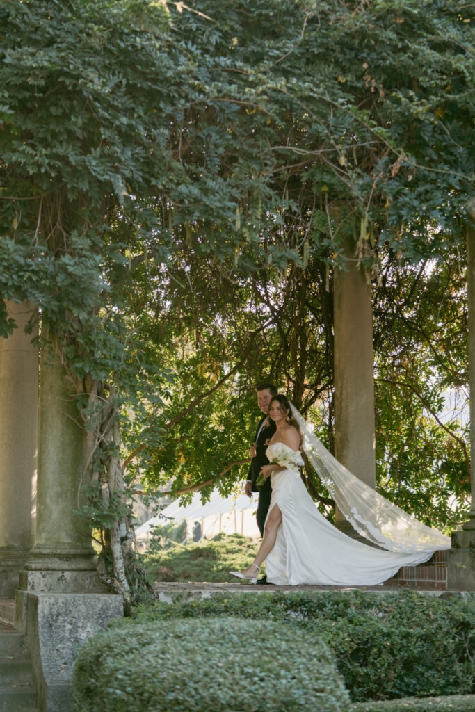 The bride walks gracefully with her escort beneath a canopy of trees and vines, her veil flowing behind her in the sunlight.