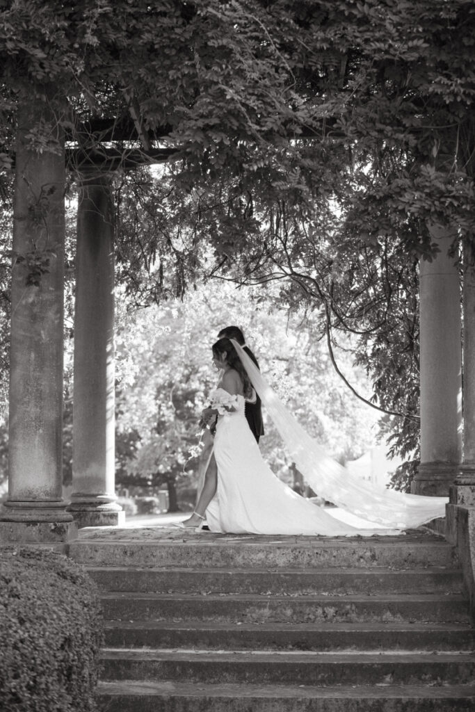 Black and white photo of bride and escort walking under vine-covered columns with sunlight in background.