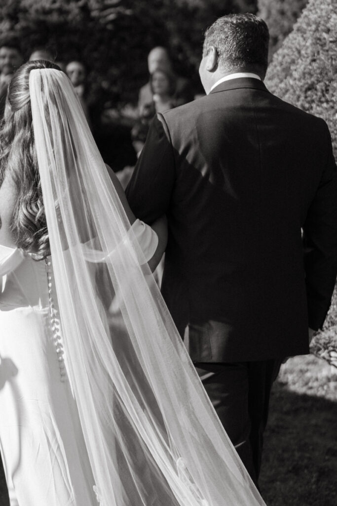 Black and white image of bride walking arm-in-arm with escort, long veil trailing behind them.