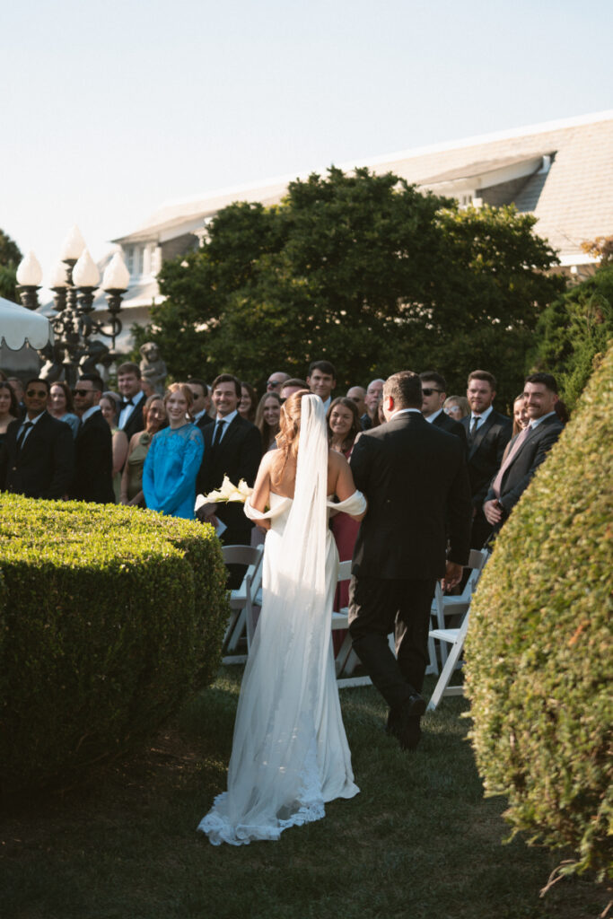 Bride walks down the outdoor wedding aisle with escort, guests smiling and watching the ceremony begin.