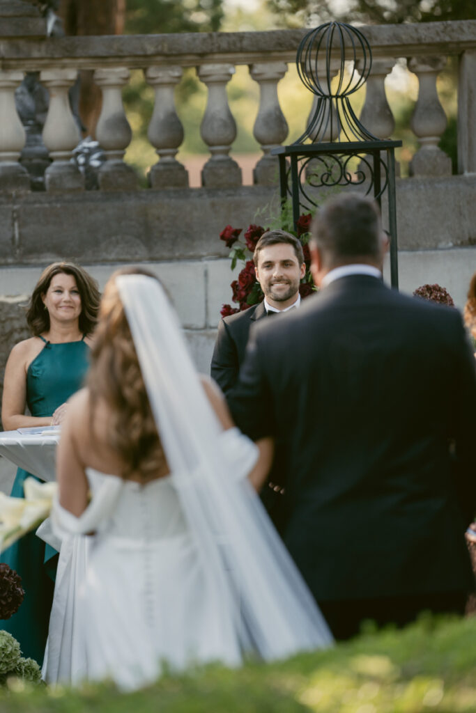 Groom smiles emotionally as bride approaches him during outdoor wedding ceremony with elegant stone backdrop.