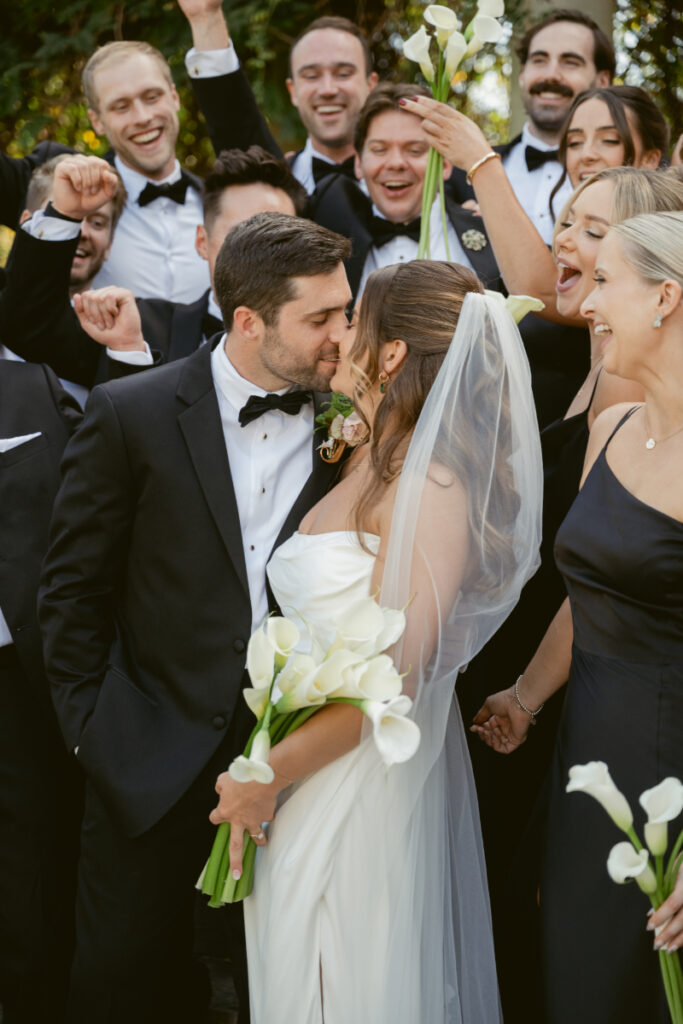 Bride and groom share a kiss with joyful bridal party cheering at Laurel Court wedding in Cincinnati.