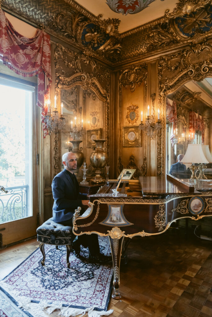 Man seated at ornate grand piano in an opulent, European-inspired room with gilded wall details.