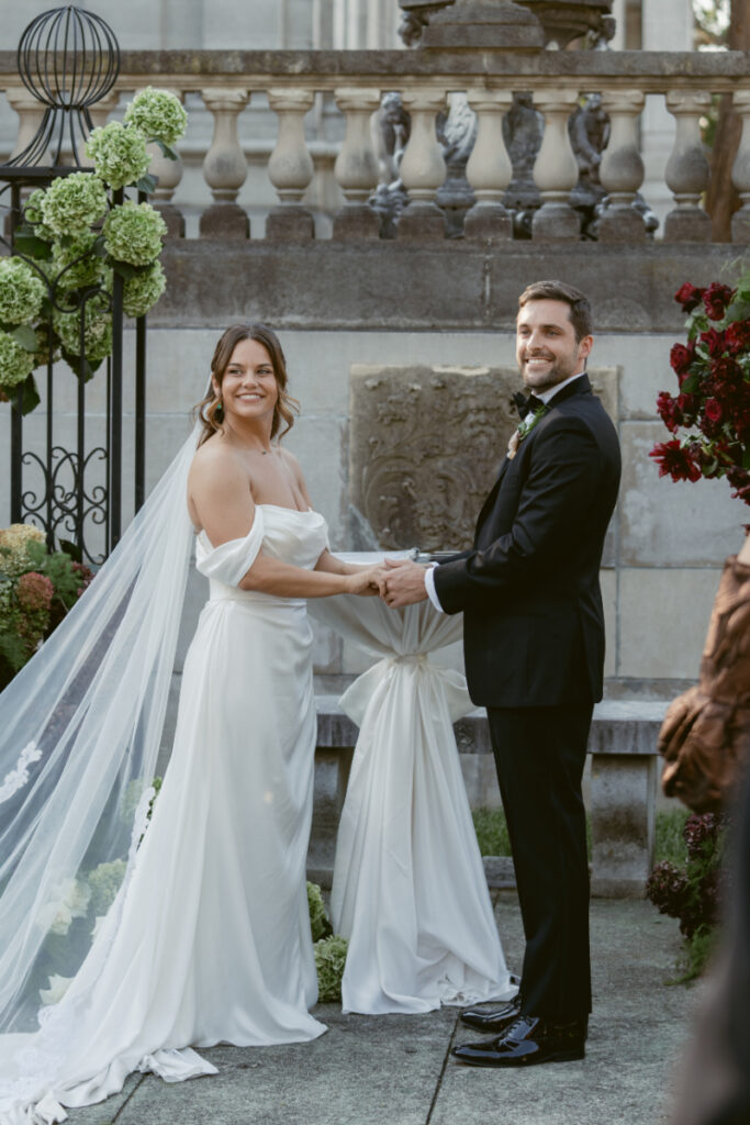 Bride and groom smile joyfully while holding hands during their elegant outdoor wedding ceremony.